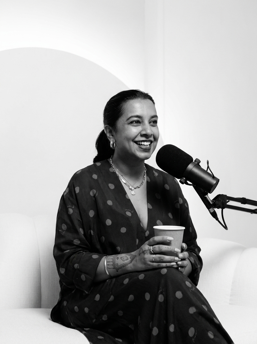A woman sitting on a white couch, smiling, holding a paper cup, with a microphone in front of her, wearing a polka dot dress and jewelry, in a studio setting.