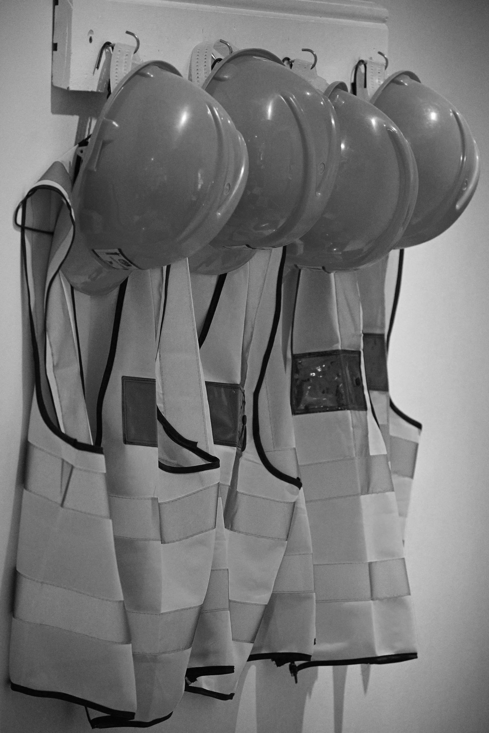 Four reflective safety helmets hanging on hooks, each with a white high-visibility vest below them, on a wall in black and white.