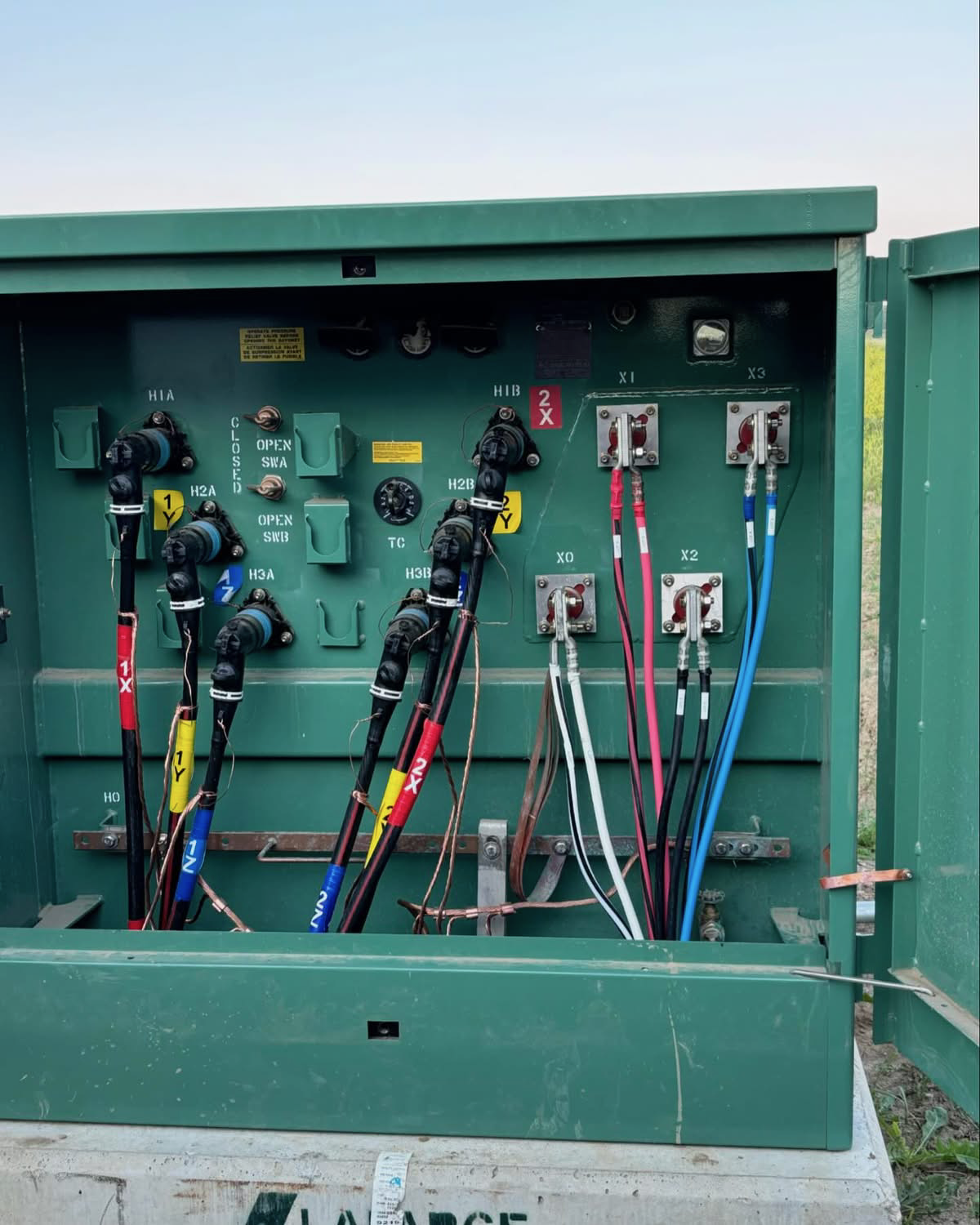 Electrical transformer with colorful wires and labeled connections, mounted on a green metal panel outdoors.