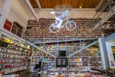 Interior of a library with shelves full of books, featuring a suspended sculpture of a person riding a bicycle. The library has a high ceiling and a modern design with a mezzanine level for additional book storage located at the LX Factory.