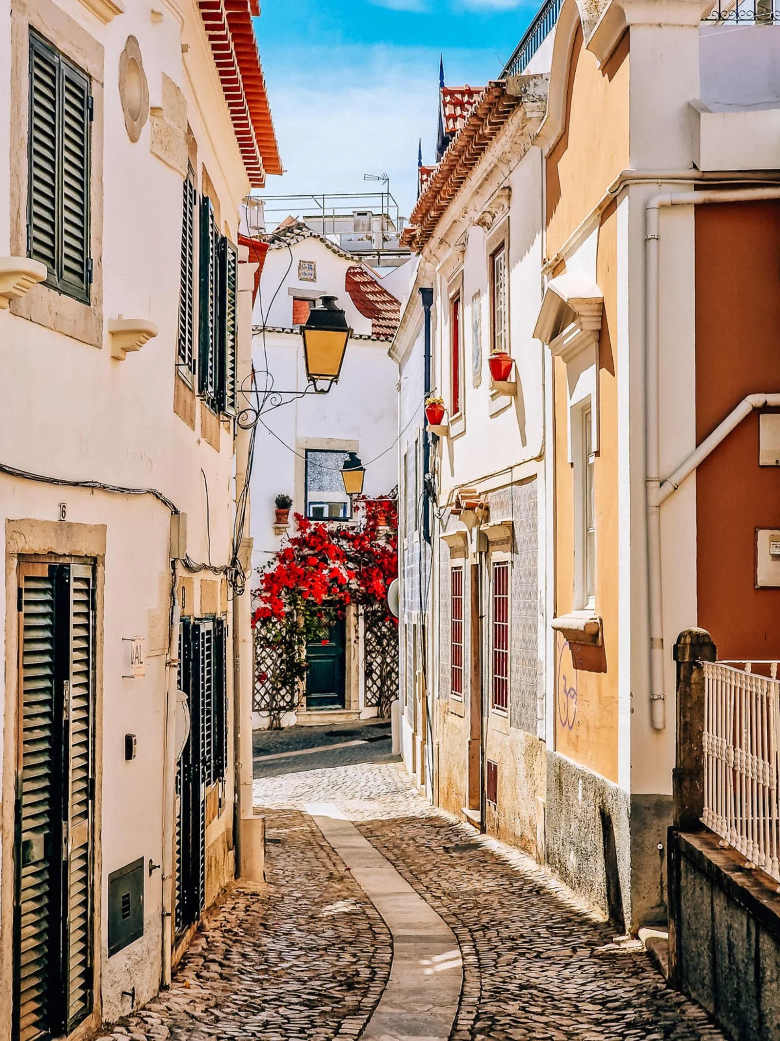 Narrow cobblestone street in historic Cascais, Portugal with white buildings adorned with red flowering plants