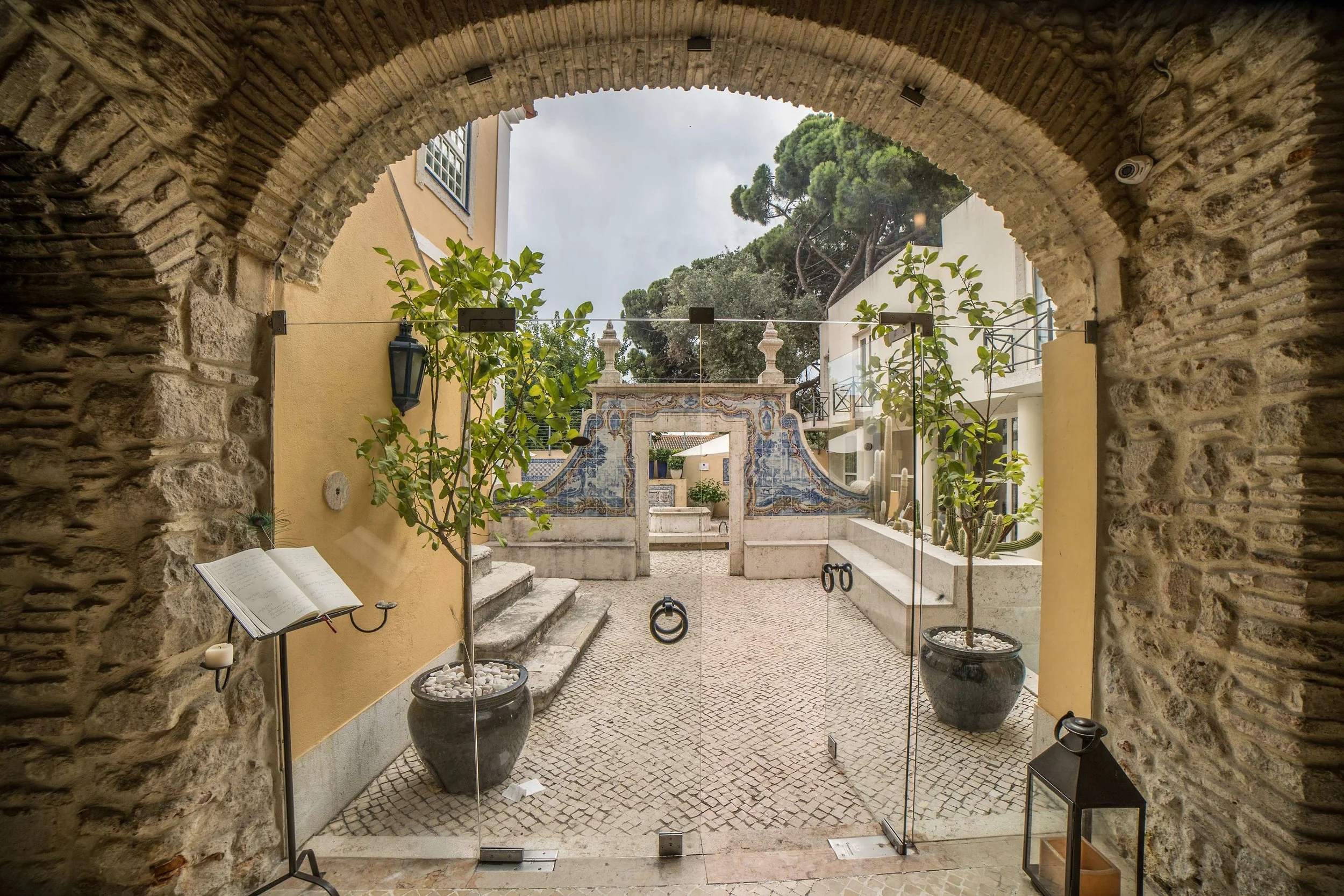 View through the arched stone entryway of the Solar do Castelo Lisbon. The archway leads to a courtyard with potted plants and a decorated fountain backdrop, featuring intricate stonework. and tile work.