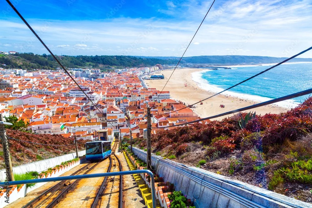 Scenic view of Nazaré, a coastal town with orange-roofed buildings, a sandy beach, and blue ocean, as seen from a funicular railway descending a hill.