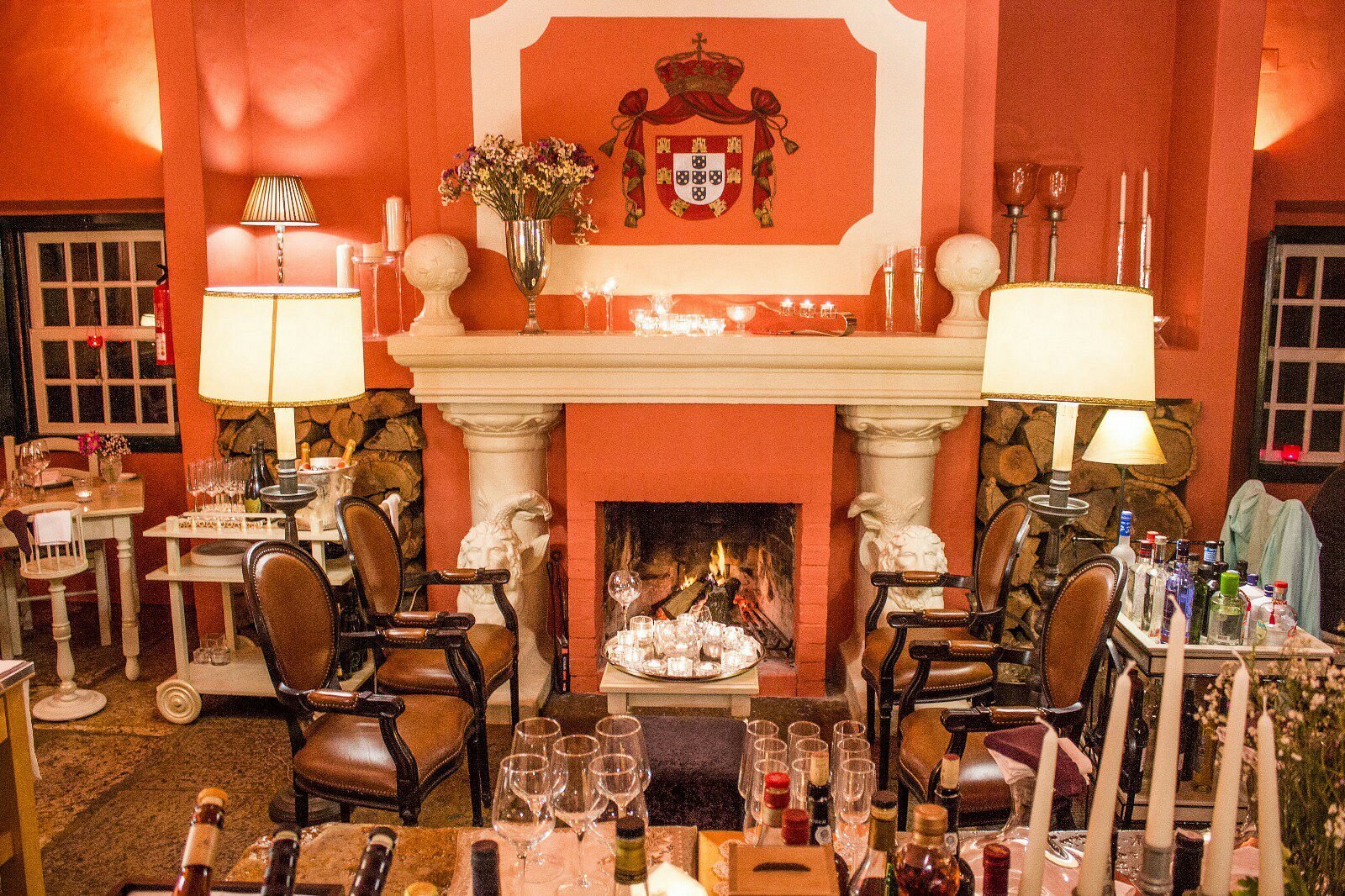 A Nova Casa de Ramiro Óbidos: Cozy Lounge with Lit Fireplace, Decorated Mantel, Armchairs, Wine Glasses, and Warm Terra-Cotta Color Scheme