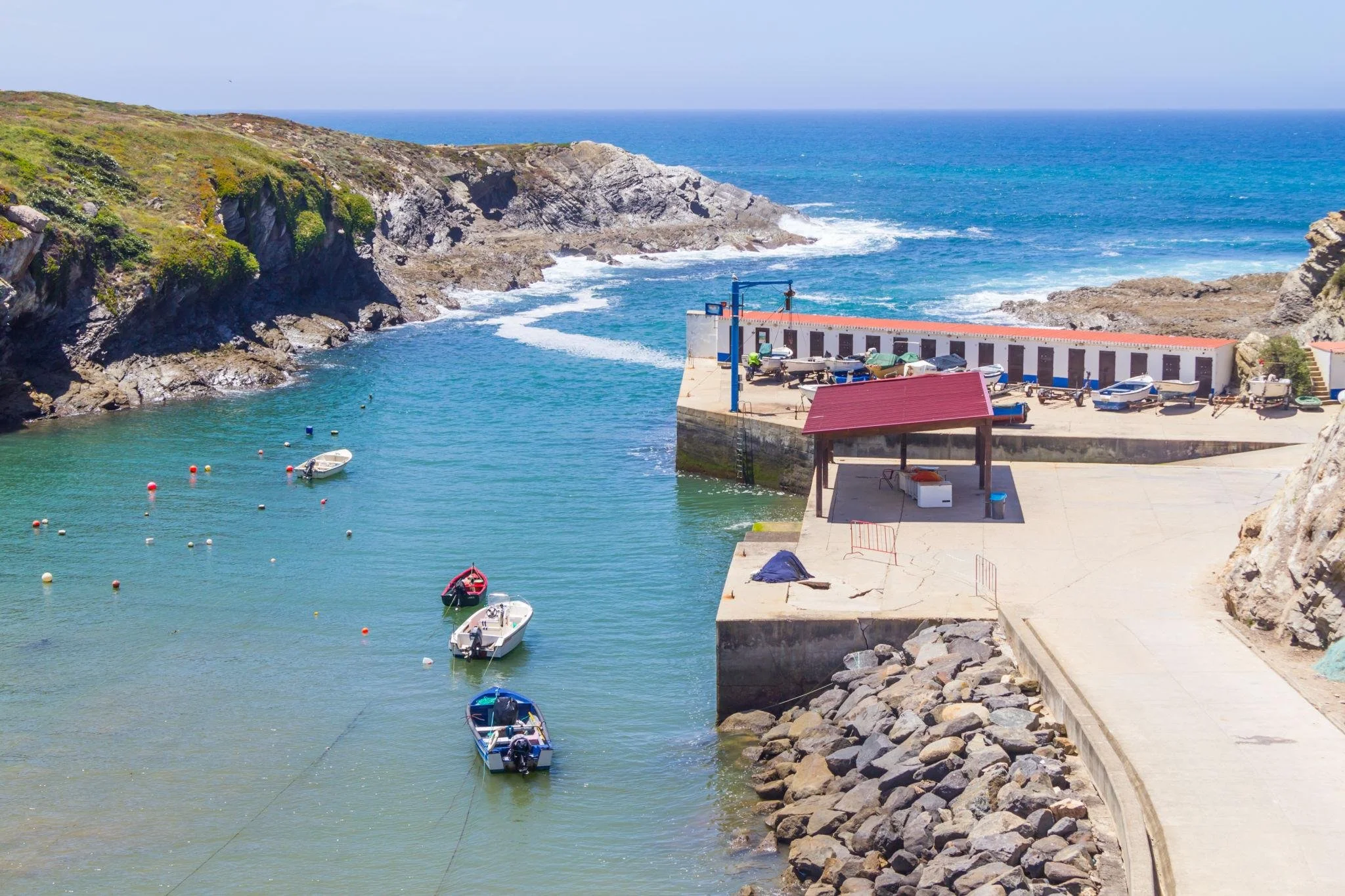 Fishing boats in a sheltered beach bay in Porto Covo, with a seaside restaurant overlooking the Atlantic Ocean.