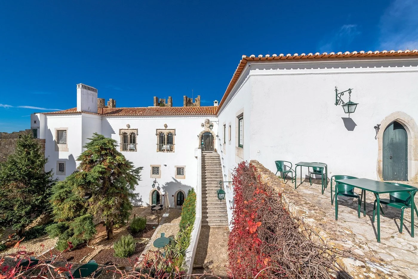 Pousada Castelo de Óbidos: Historic White-Facade Building with Exterior Staircase, Garden, and Green Tables on a Stone Terrace.