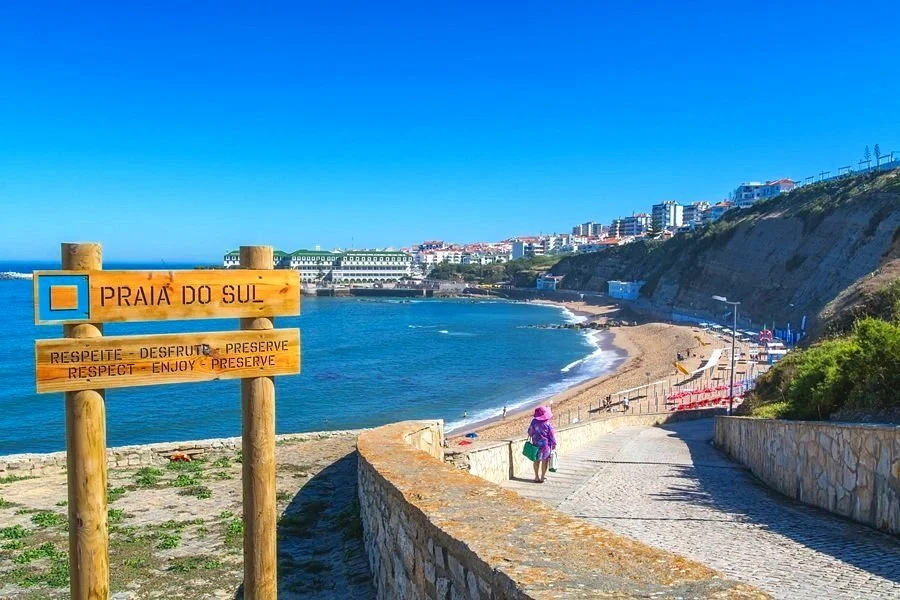 Praia do Sul Beach in Ericeira, Portugal: Scenic view with a beach sign, a woman walking down the path alongside the cliffs, down to the ocean, with coastal buildings in the background