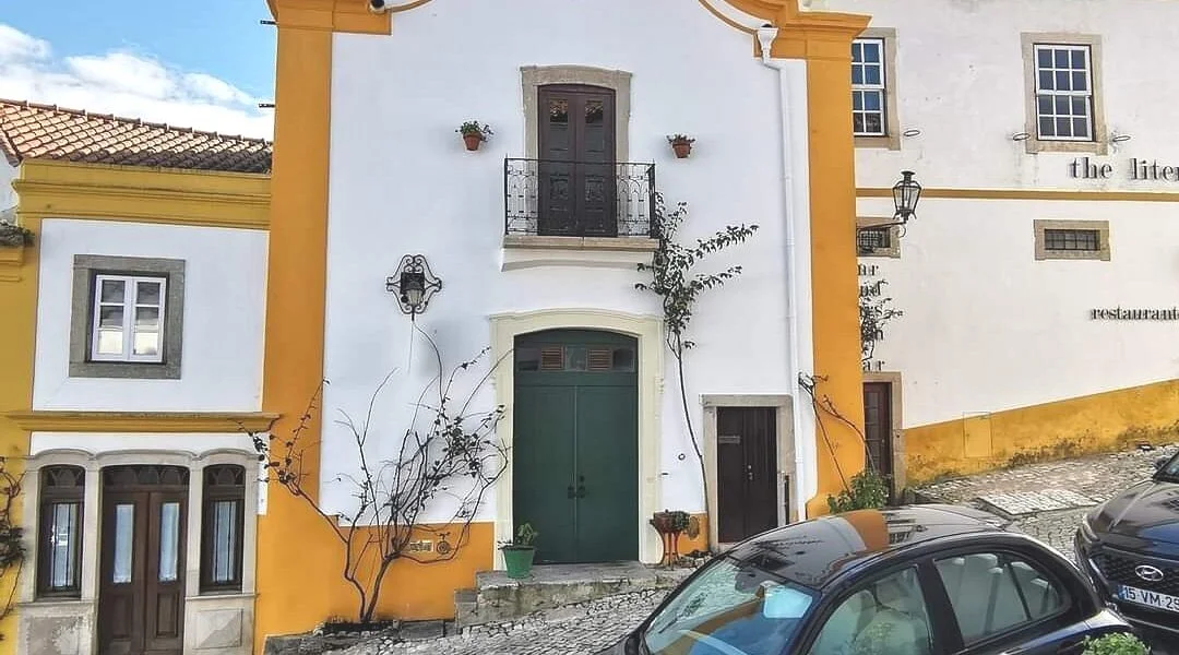 Pontinha Restaurant, Óbidos: Charming White and Yellow Building with Small Balcony, Green Door, Potted Plants, and Cobblestone Street.
