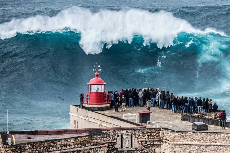 Large ocean wave approaching a coastal rooftop with a crowd of people and a red lighthouse in Nazare.