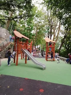Jardins do Palácio de Cristal, Porto: Children's Playground with Wooden Structures, Slide, and People Enjoying the Tree-lined Area.