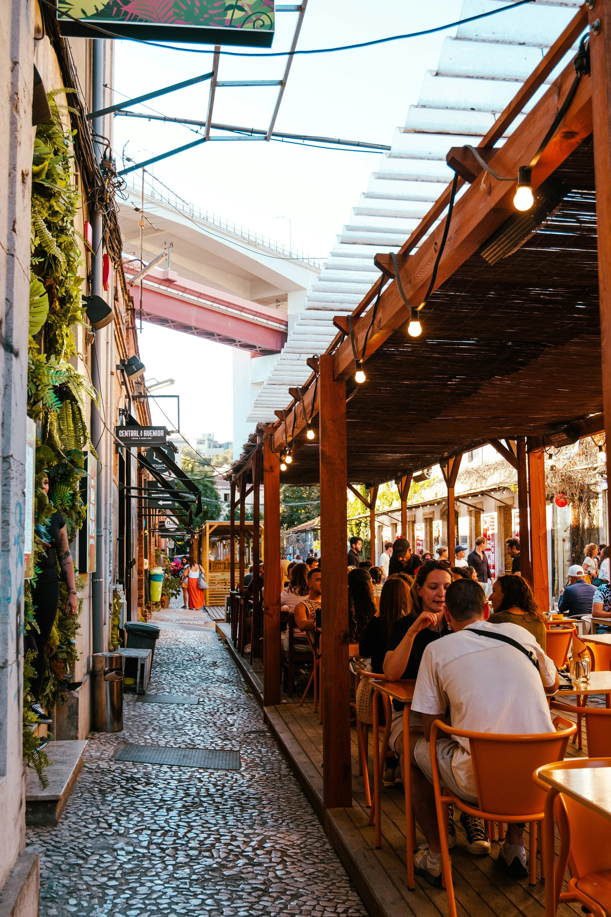 Outdoor cafe at LX Factory Lisbon with people dining, cobblestone pathway, string lights, and overhead bridge.