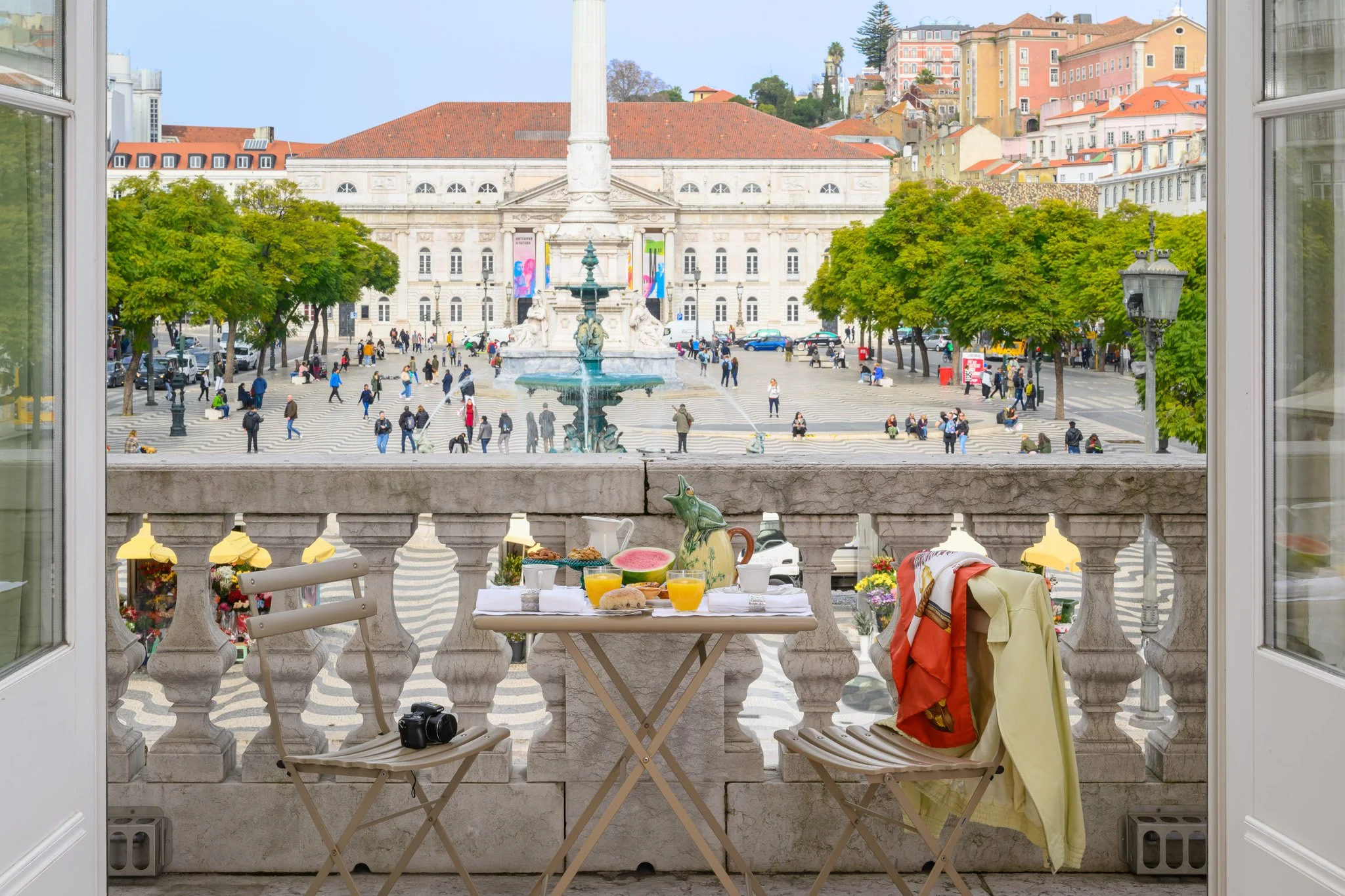 View from a balcony of the Residentas Arco do Bandeira hotel, overlooking a fountain in the public square, surrounded by classical Lisbon architecture. A table is set with breakfast items, including fruit and orange juice.