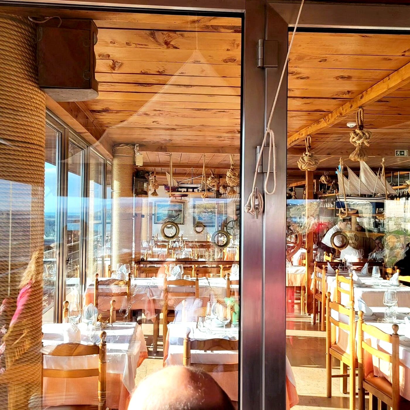 Interior of Esplanada Furnas, Ericeira: Nautical-Themed Restaurant with Wooden Furniture, White Tablecloths, Glassware, Ship Models, and Large Windows.