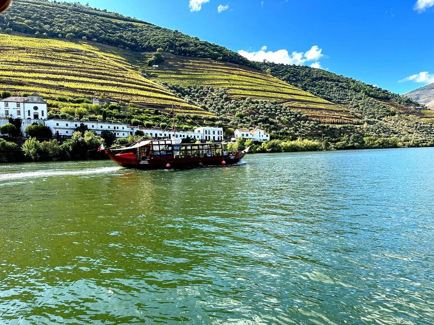 Scenic view of a traditional boat on the Douro River with terraced vineyards and houses in the background, under a bright blue sky.