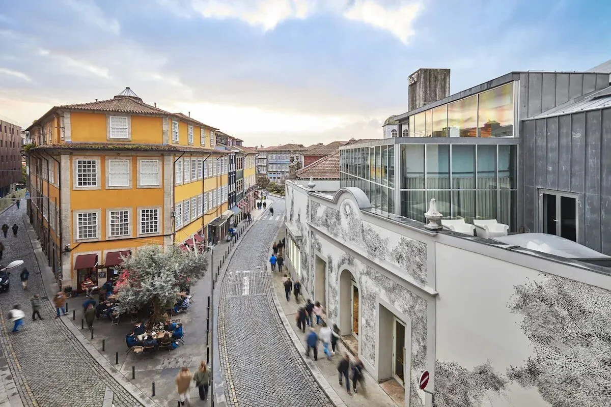 Yellow corner building and cobbled street in Porto's Baixa, with cafe tables under a tree, an azulejo-tiled facade and a modern glass extension alongside.