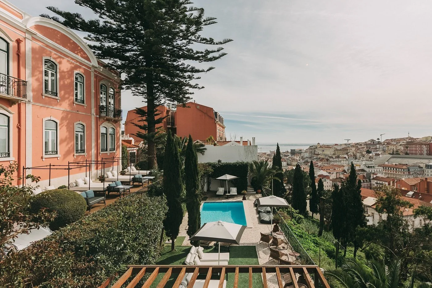View of the Torel Palace, with a terra-cotta-colored exterior lined with balconies overlooking the Tagus River and swimming pool, with the Lisbon cityscape in the background.