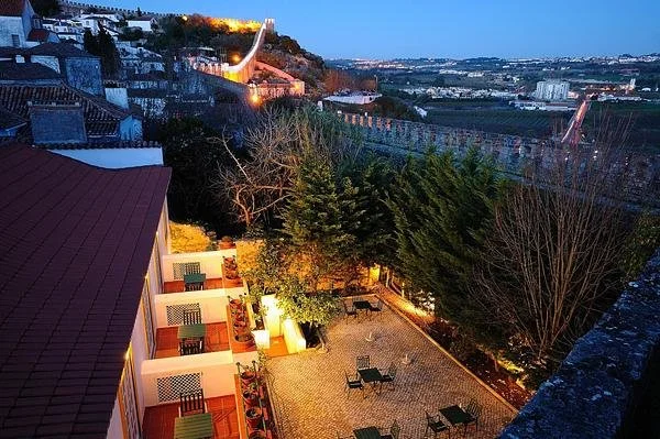 Scenic view of Casa Senhoras Rainhas, Obidos, Portugal at dusk with illuminated medieval walls, stone pathways, and outdoor seating areas.