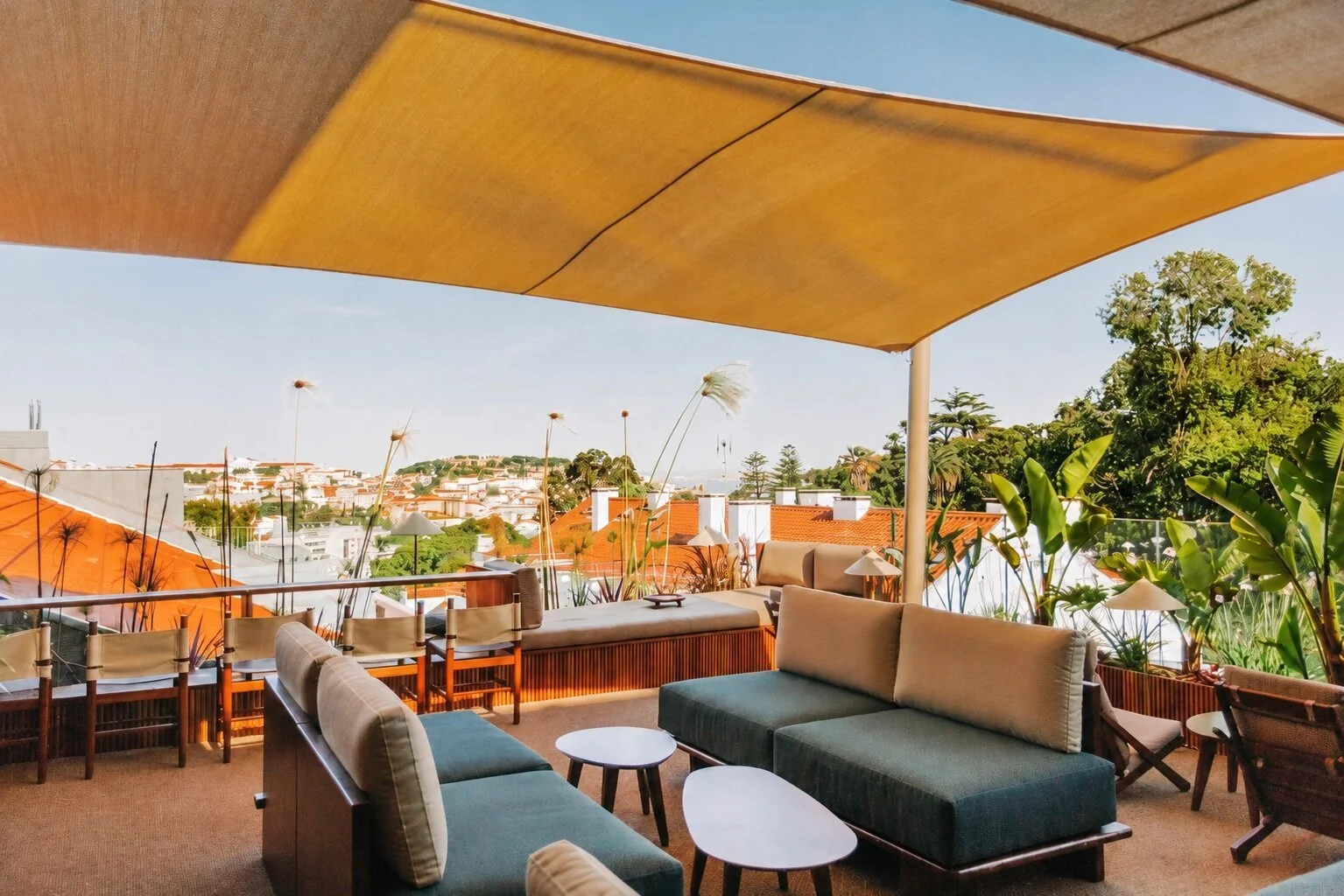 Shaded rooftop terrace in Lisbon with lounge sofas and chairs beneath a canvas canopy, looking out over the city's rooftops and towards the castle hill.