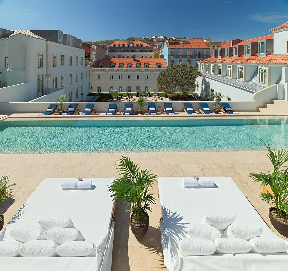 View of the interior pool area with lounge chairs at the elegant One Palácio da Anunciada. The pool looks over a landscaped courtyard with whitewashed buildings and red-tiled roofs in the background.