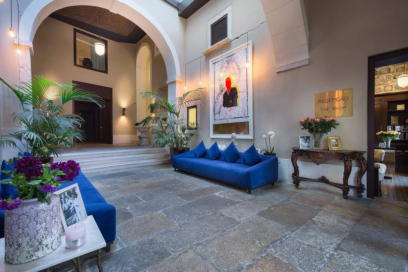 The luxurious hotel lobby of the Santiago de Alfama Lisbon with blue velvet couches, tropical plants, modern artwork, and warm lighting. Features stone flooring and elegant decor.