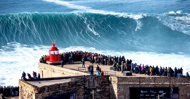Crowd watching surfers riding a massive wave at a coastal viewpoint in Nazaré.