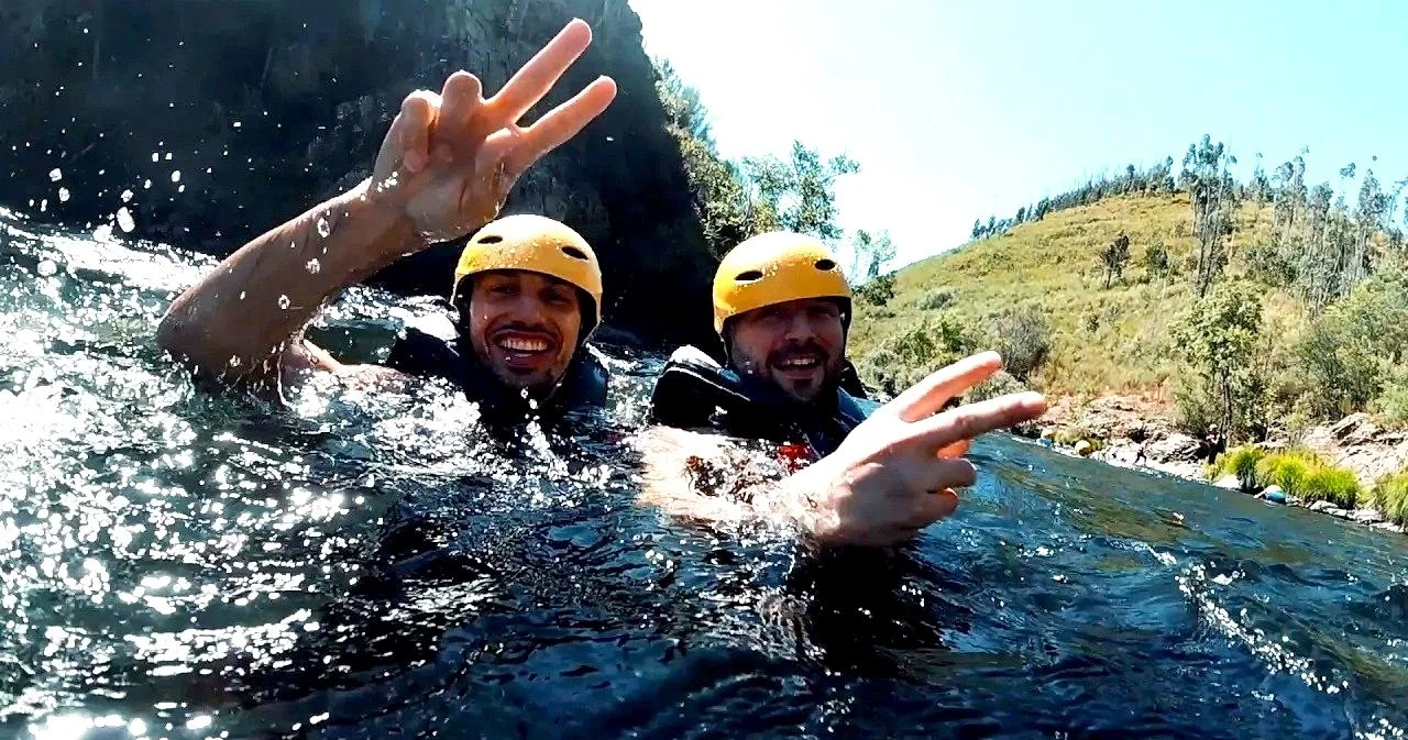 Two people wearing yellow helmets and life jackets enjoying river trekking on the Paiva river, making peace signs.