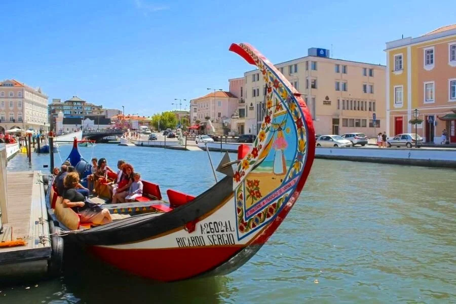 Colorful Moliceiro boat with passengers on a canal in Aveiro, Portugal. Buildings and blue sky can be seen in the background.