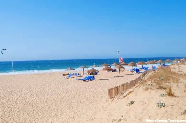 Foz do Lizandro Beach in Ericeira: Sandy Shores with Straw Umbrellas, Blue Lounge Chairs, and Clear Blue Ocean.