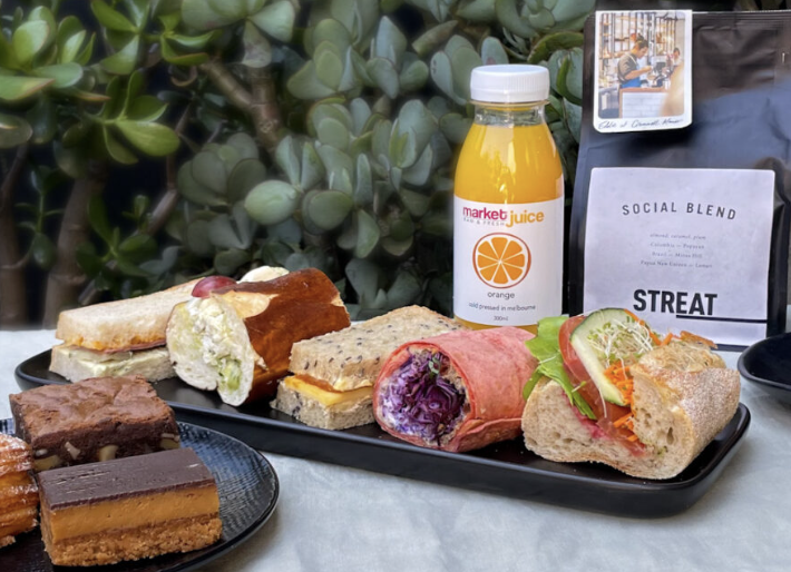 a variety of slices, cakes, sandwiches and a juice on a table with a bag of coffee beans displayed behind