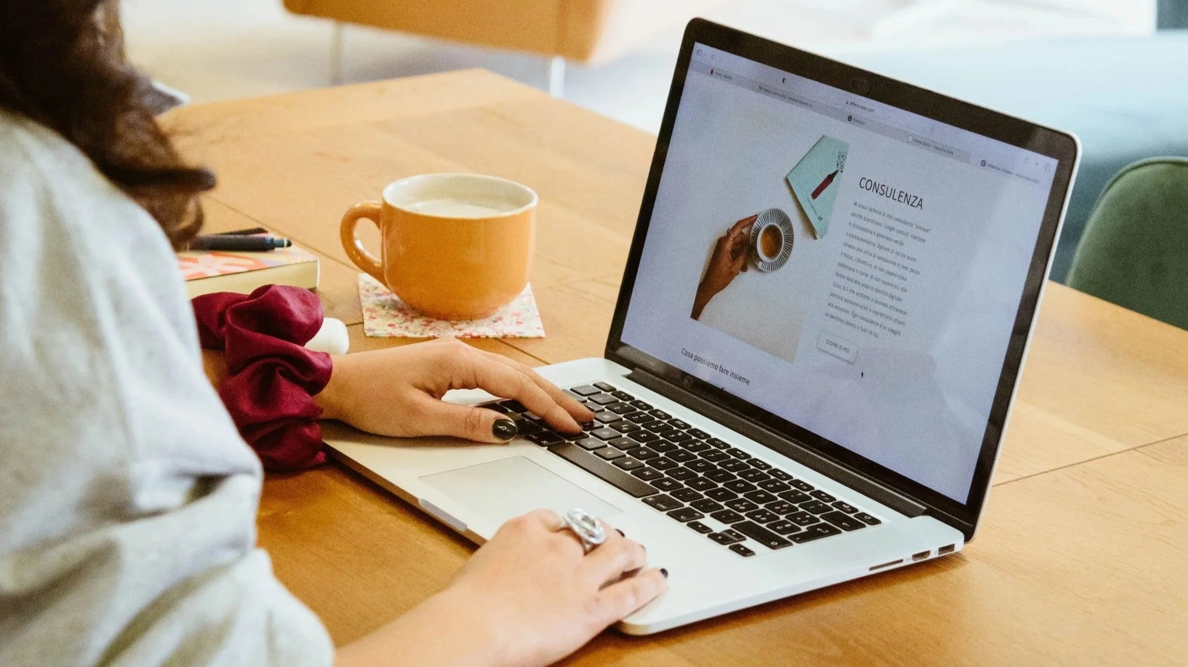 Persona che utilizza un laptop sul tavolo, con una tazza arancione di caffè, un quaderno e una penna accanto.