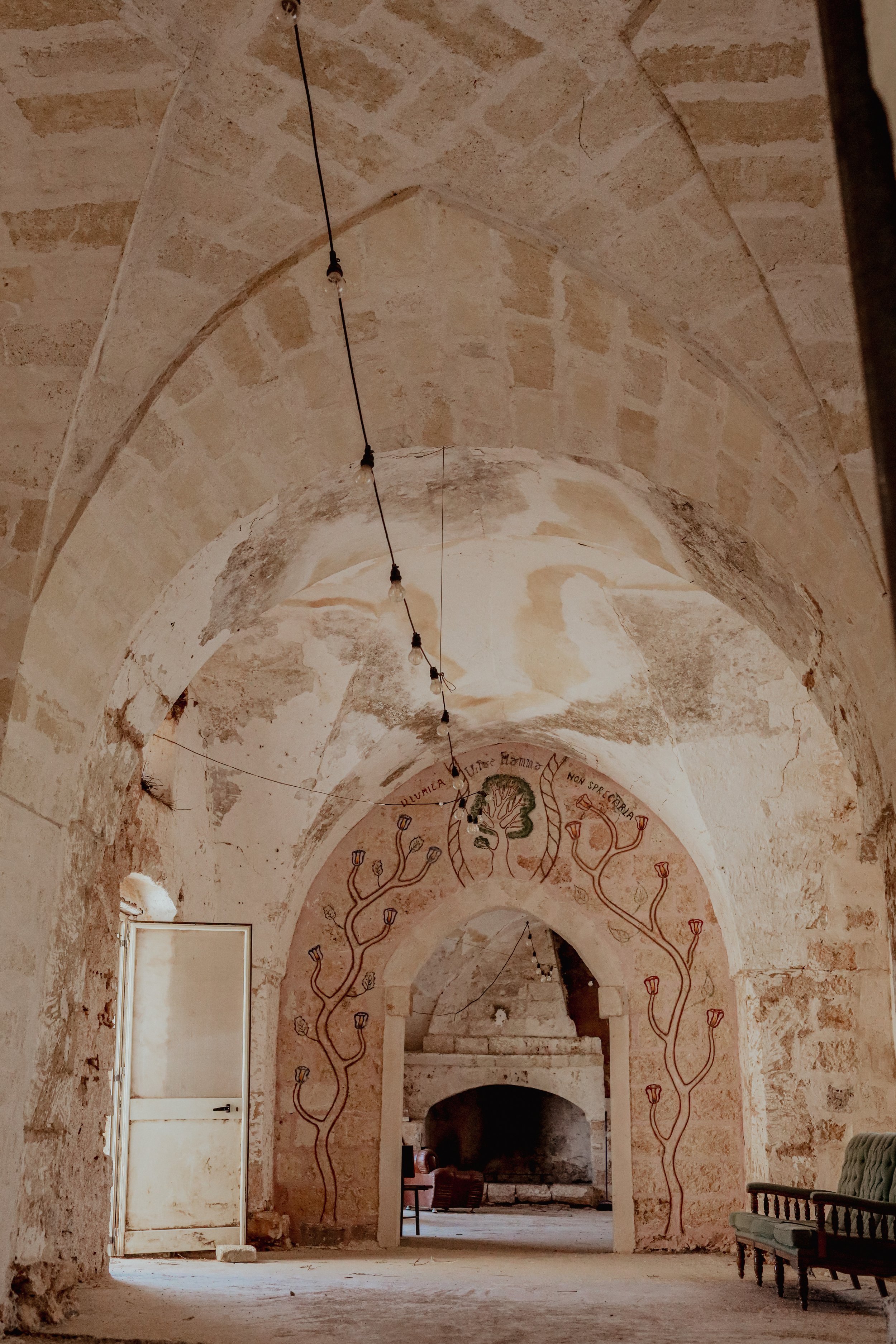 Interno di una stanza con archi in pietra, decorata con un disegno di alberi stilizzati e scritte in latino sopra l'arco, lampade a sospensione, pareti e soffitto in pietra.