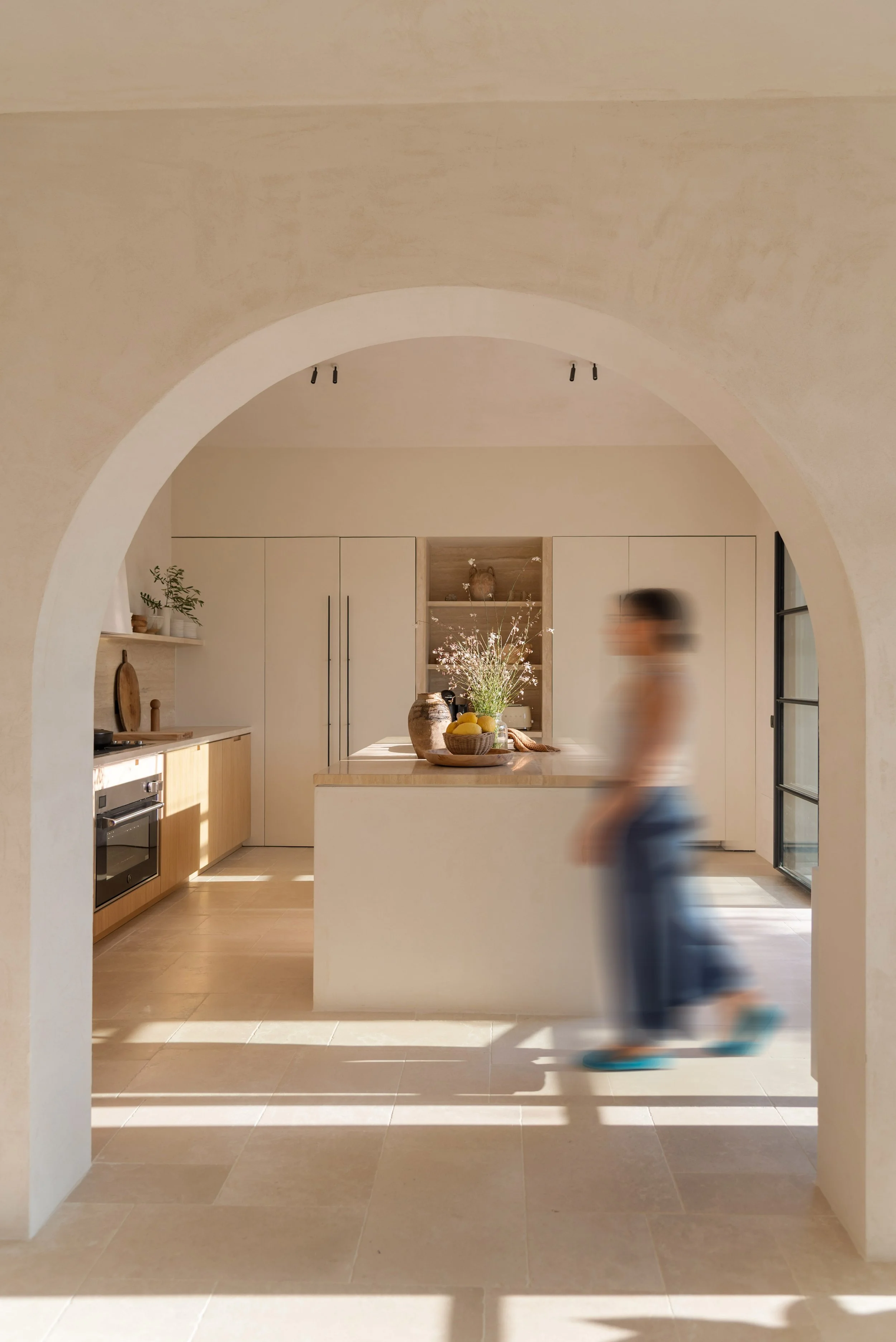 Interno di una cucina moderna vista attraverso un arco, con un'apertura sulla sala, con un piano cucina centrale decorato con vasi e fiori, armadietti bianchi e una finestra grande che lascia entrare molta luce naturale, e una donna che cammina velocemente nell'immagine.