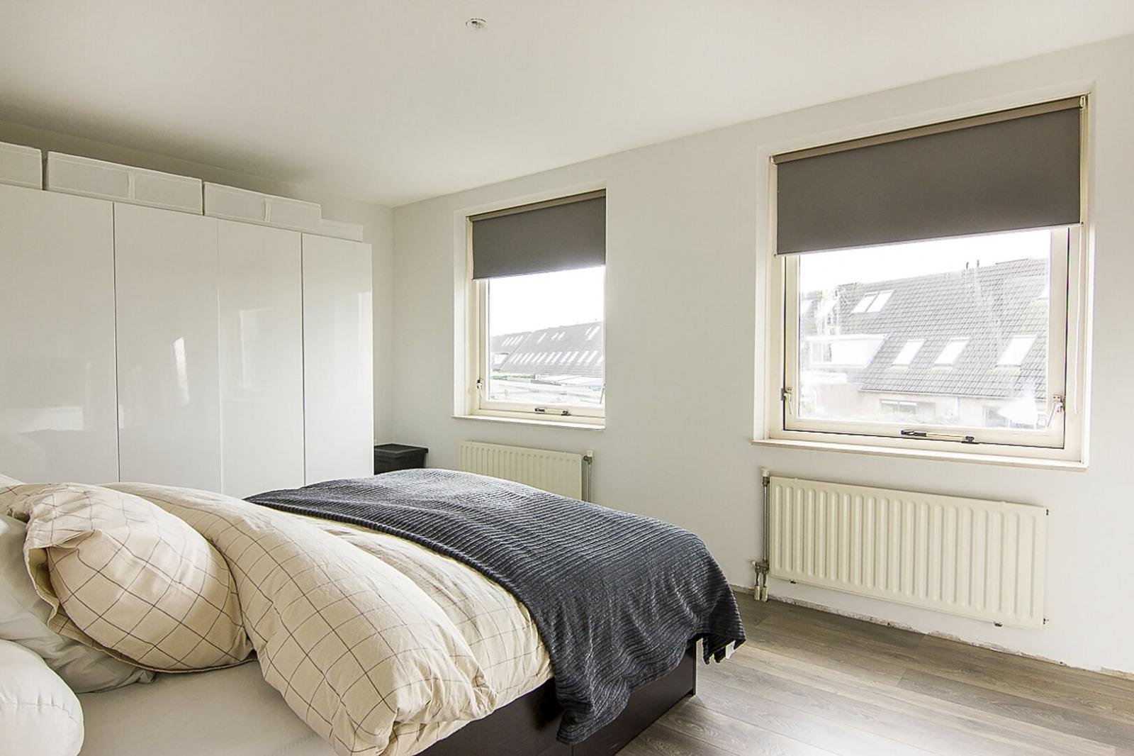 Slaapkamer met wit bed, beige kussens, donkergrijze deken, witte kasten, twee ramen met donkere rolgordijnen, houten vloer, radiatoren onder ramen, uitzicht op huizen met daken