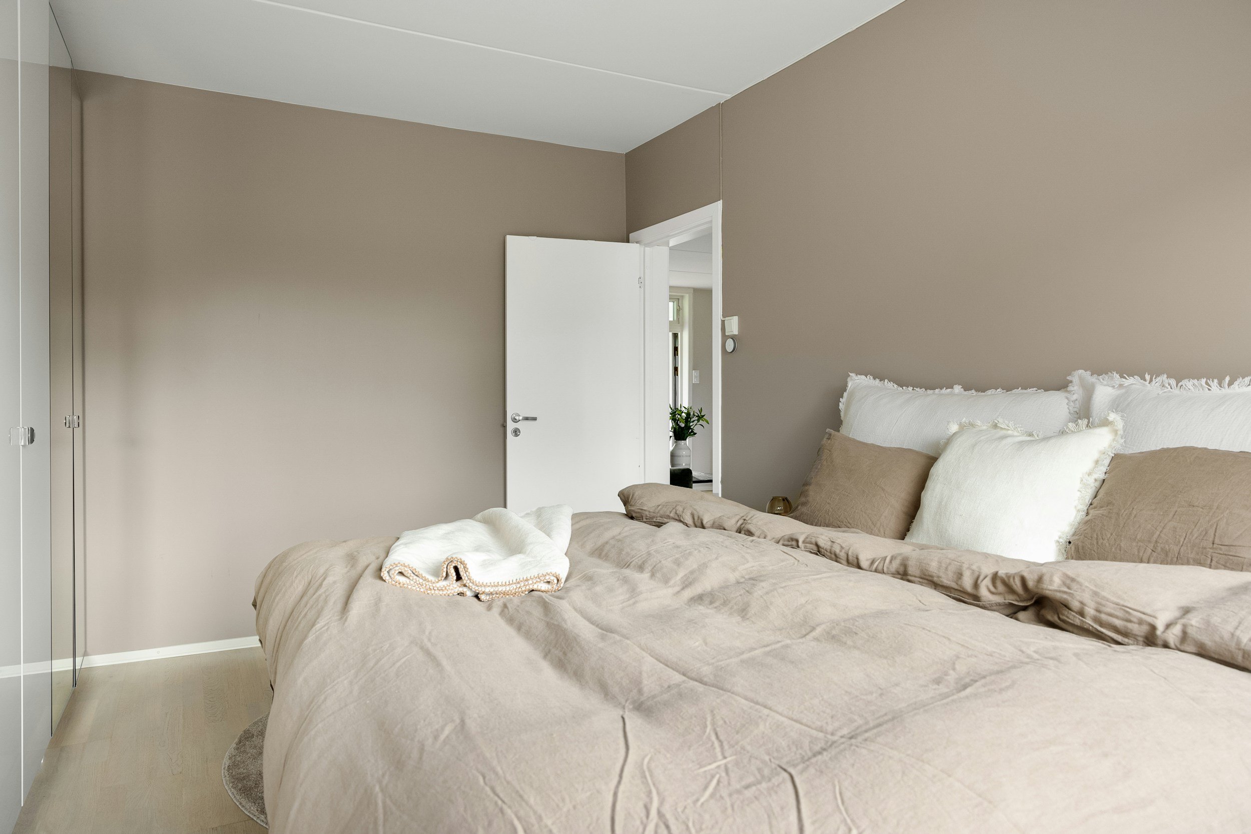 Een slaapkamer met beige beddengoed en witte en beige kussens, een open witte deur, een plant in de deuropening, beige muren en een witte plafond.