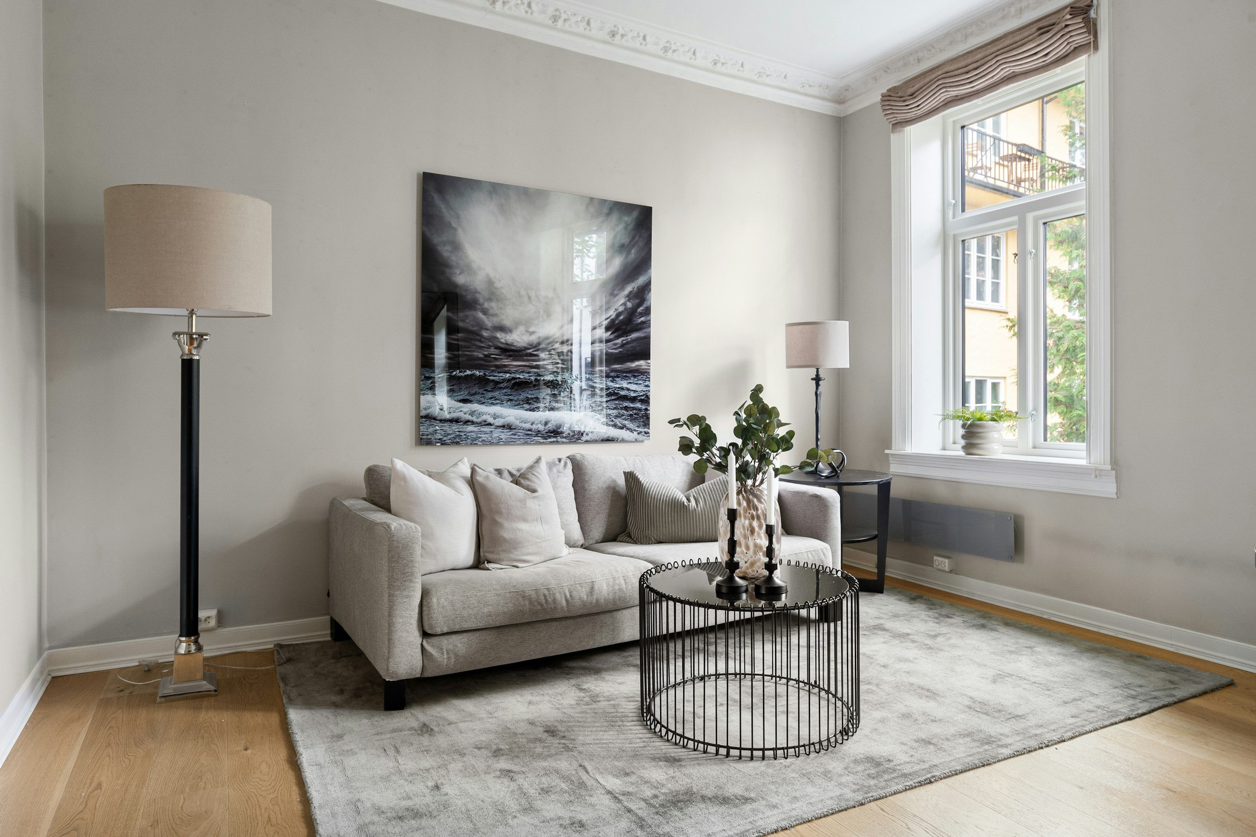 Woonkamer met een lichtgrijze bank, een ronde zwarte tafel met bloemen, een grote muurkunstwerk van de zee, twee lampen met beige lampenkappen, een grote raam met witte kozijnen, en een grijze tapijt op houten vloer.