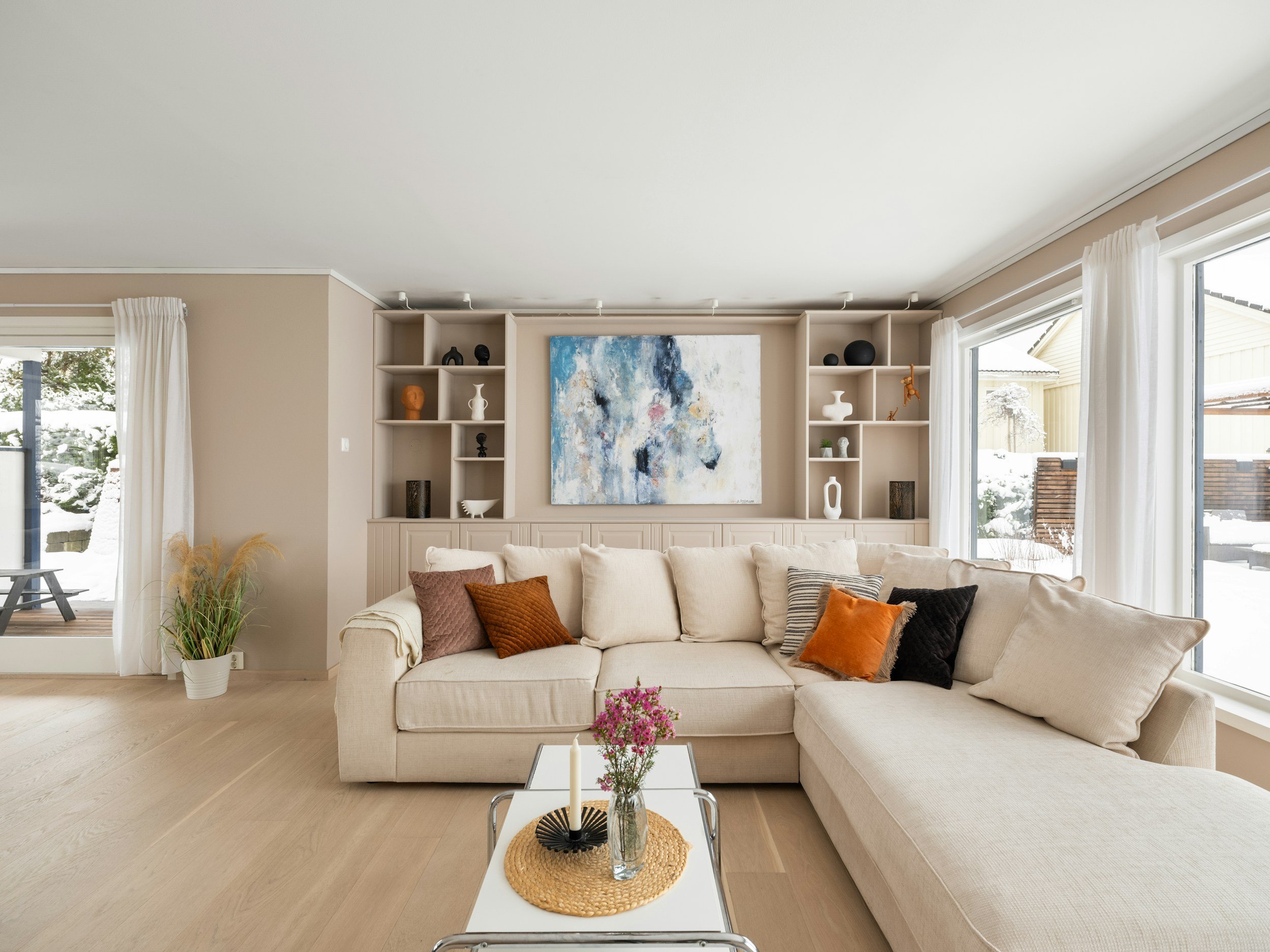 Lichte woonkamer met beige bank en kussens, witte boekenkast met decoratieve objecten, abstract schilderij aan de muur, grote ramen met witte gordijnen, houten vloer en een koffietafel met bloemen.