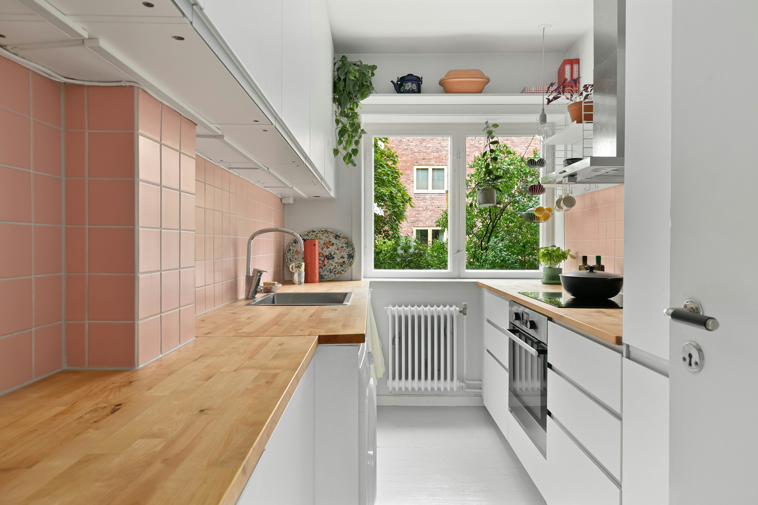 Een moderne, lichte keuken met witte kasten, houten werkbladen en een grote ramen met uitzicht op groene bomen en een gebouw.