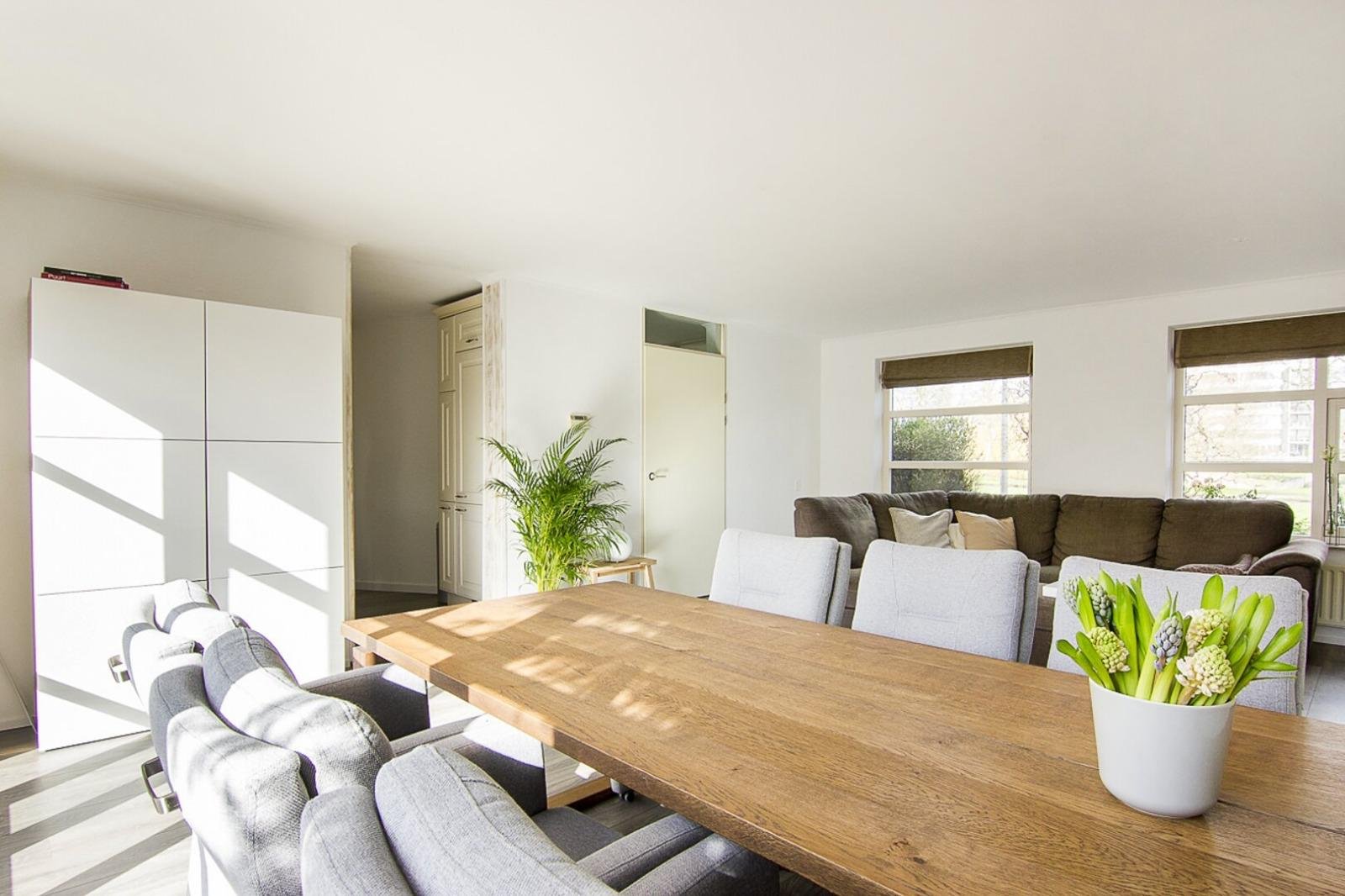 Een woonkamer met een grote houten eettafel, grijze eetstoelen, een grote bank, een groene plant, en grote ramen met donkere jaloezieën.