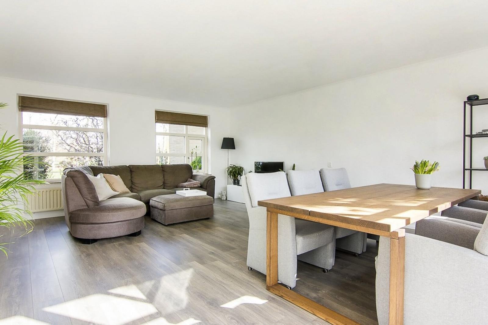 Lichte woonkamer met grote ramen, een grijze hoekbank, een houten eettafel met lichtgrijze stoelen, en planten.
