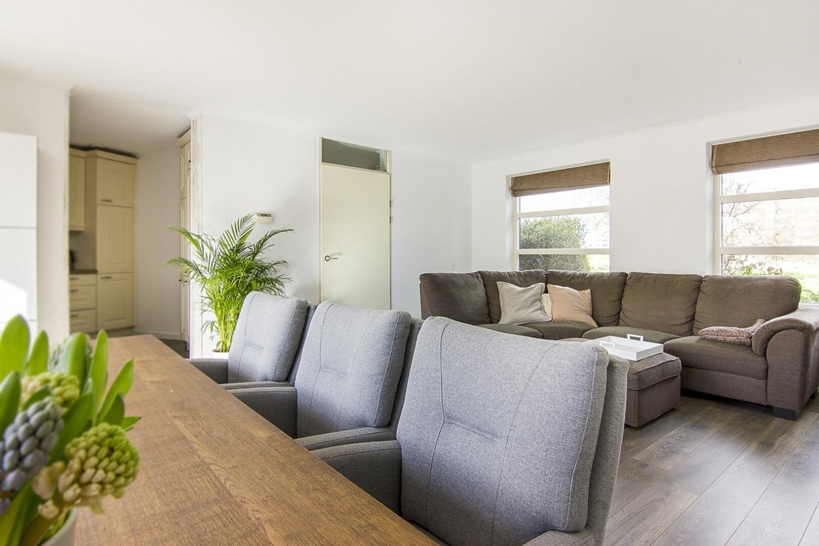 Woonkamer met grote L-vormige bank, salontafel, grote ramen, houten vloer, en planten.