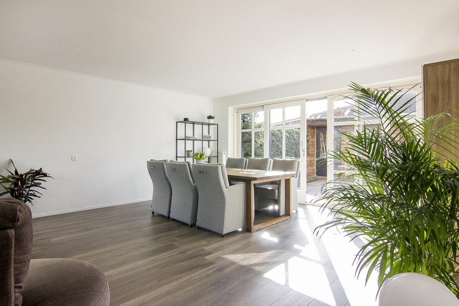 Ruim, modern woon- en eetkamer met grote ramen, witte muren, houten vloer, en een grote planten