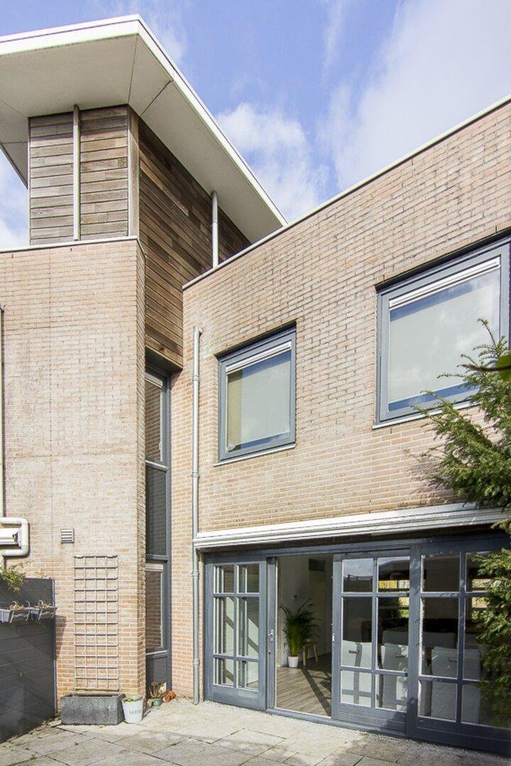 Modern stenen woning met grote glazen deuren en ramen, tuin met planten, onder een blauwe hemel.