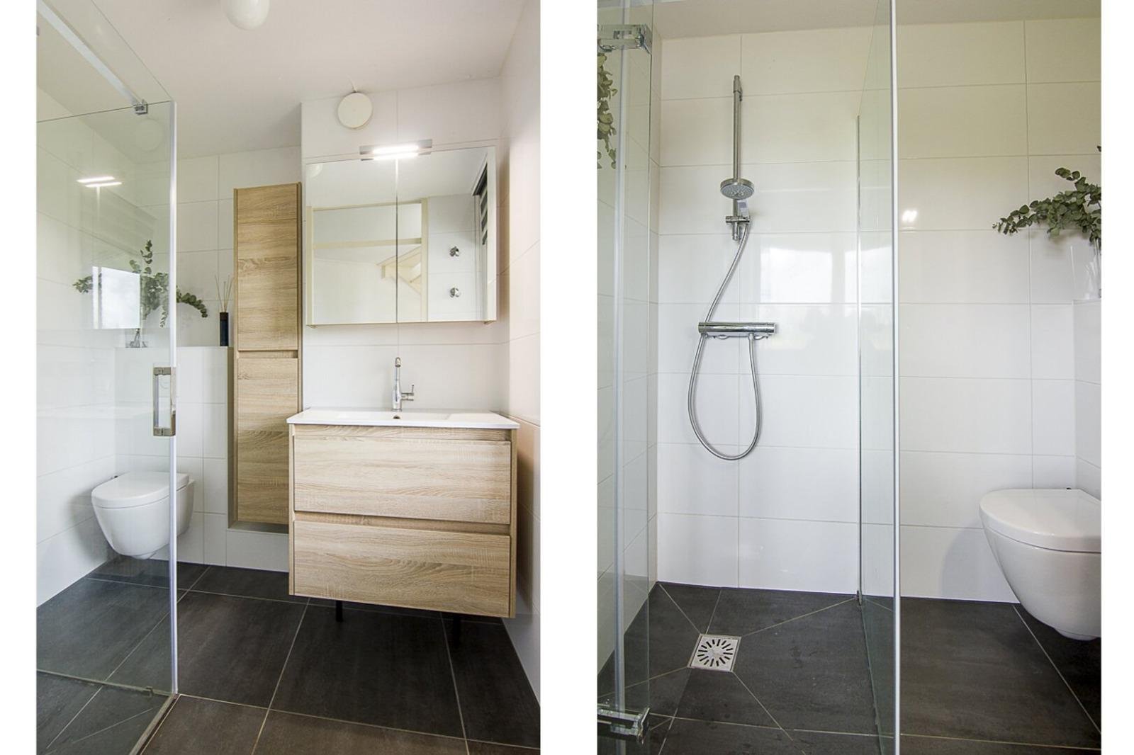 Een moderne badkamer met een inloopdouche, een zwevend toilet en een houten wastafel met spiegelkast. Gedeeltelijk door een glazen wand gescheiden, lichte muur en donkere vloeren.