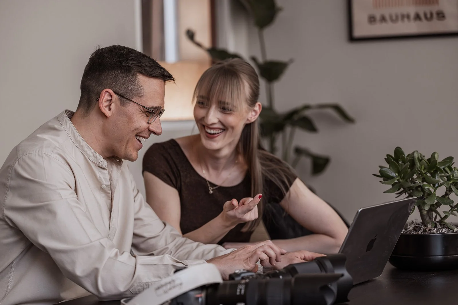 Zwei lächelnde Menschen, ein Mann mit Brille und eine Frau, sitzen zusammen an einem Tisch mit einem Laptop und einem Videokamera, lachen und unterhalten sich, im Hintergrund Pflanzen und eine Wand mit einem Gemälde, in einem gemütlichen Raum.
