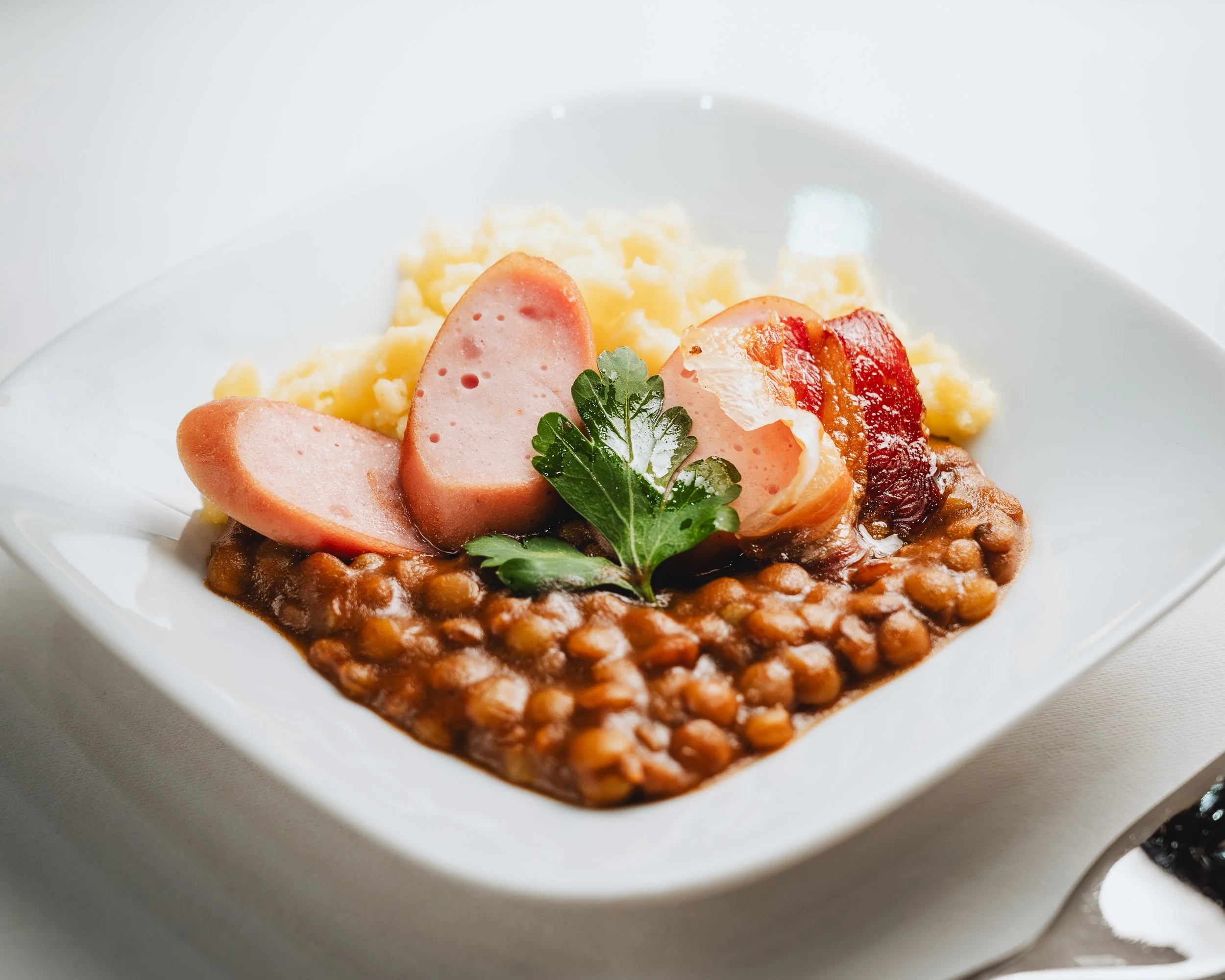 Ein Teller mit Linsensuppe, gekochtem Kartoffelpüree, Scheiben von Wurst, Bacon, und frischer Petersilie.