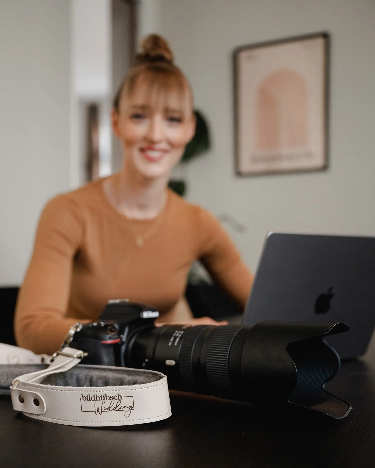 Eine lachende Frau sitzt an einem Tisch mit Kamera, Laptop und Fotografie-Ausrüstung, im Hintergrund eine Pflanze und ein Wandbild.