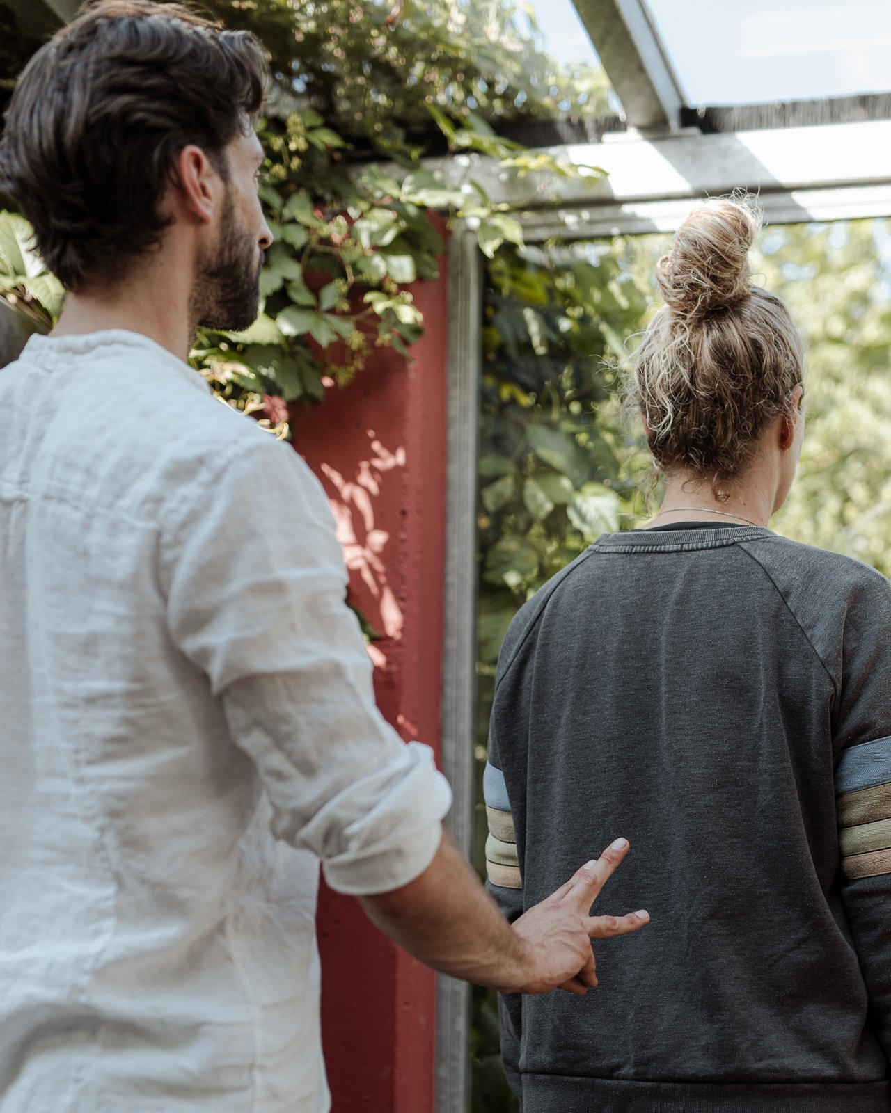 Ein Mann mit dunklen Haaren und Bart zeigt mit seinem Finger auf den Rücken einer Frau mit blondem, lockigem Haar, das zu einem hohen Dutt zusammengebunden ist, in einem Gewächshaus. Im Hintergrund sind grüne Pflanzen und Sonnenlicht sichtbar.