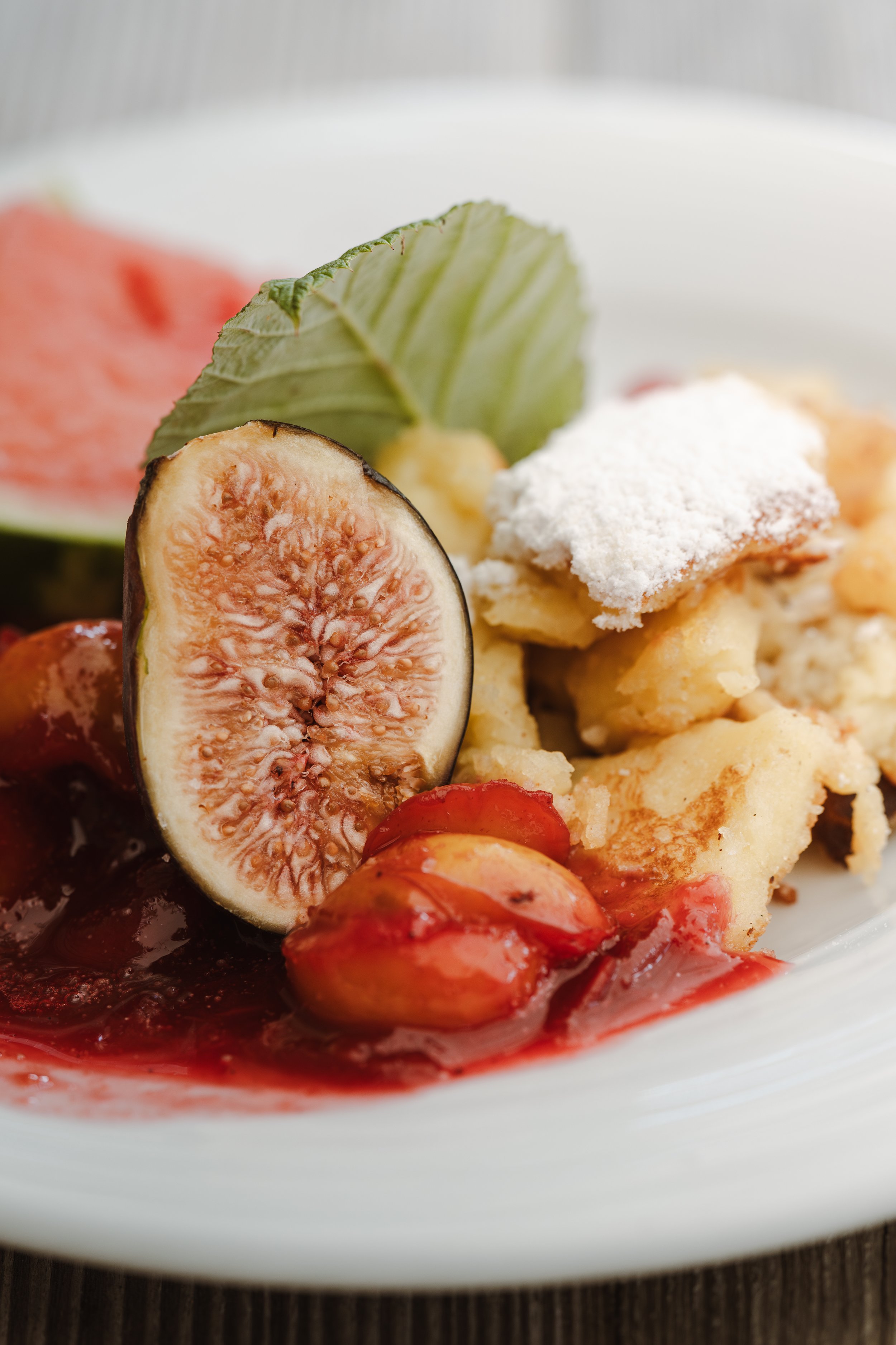 Ein Teller mit Fruchtkompott, frischen Feigen, Erdbeeren, Sahne, einer grünen Weinblatt und einem Stück Wassermelone.