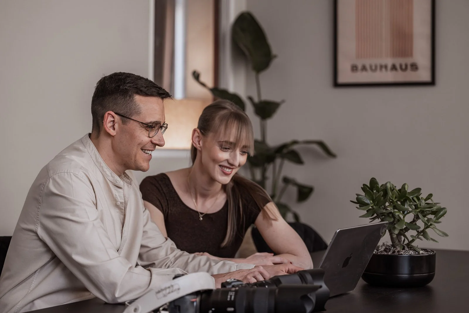 Ein Mann und eine Frau schauen gemeinsam auf einen Laptop, beide lächeln. Auf dem Tisch liegt eine Kamera und ein Telefon. Im Hintergrund ein Wandbild mit dem Wort 'BAUHAUS' und eine Pflanze.