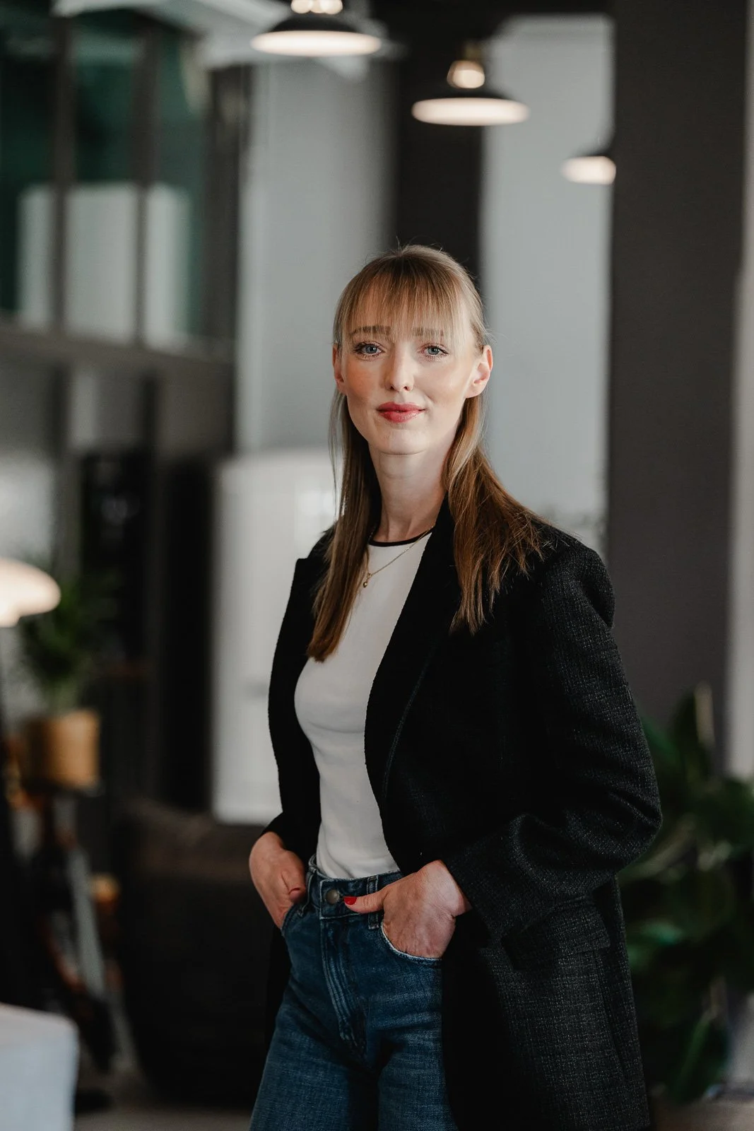 Business Shooting im Studio – Personal Branding Fotografie für LinkedIn - Eine junge Frau in einem schwarzen Blazer und blauen Jeans steht in einem modernen Raum mit Bürolampen im Hintergrund.